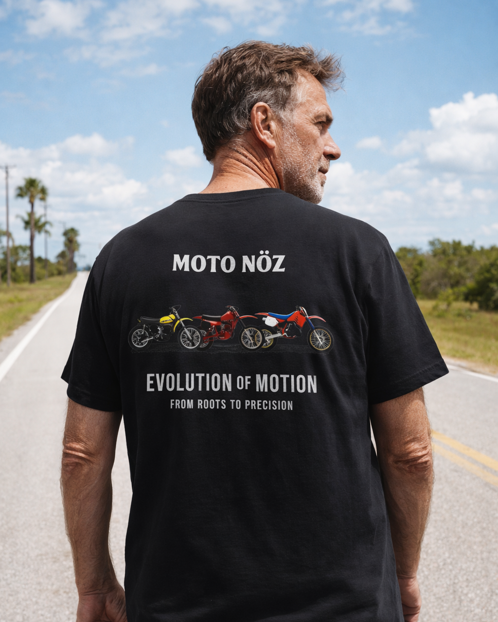 Man wearing a black t-shirt with 'Moto Noz' design on a road with a clear sky.