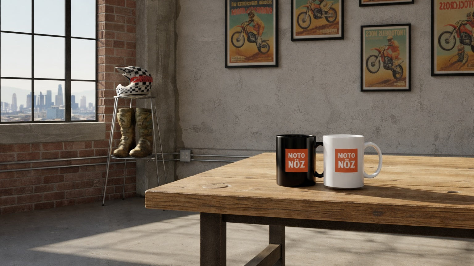 Two mugs on a wooden table with posters of motorcyclists on the wall in a room with large windows and brick wall.